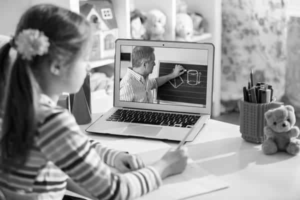 A young student focused on her laptop screen during an online class, taking notes with a pencil and paper.