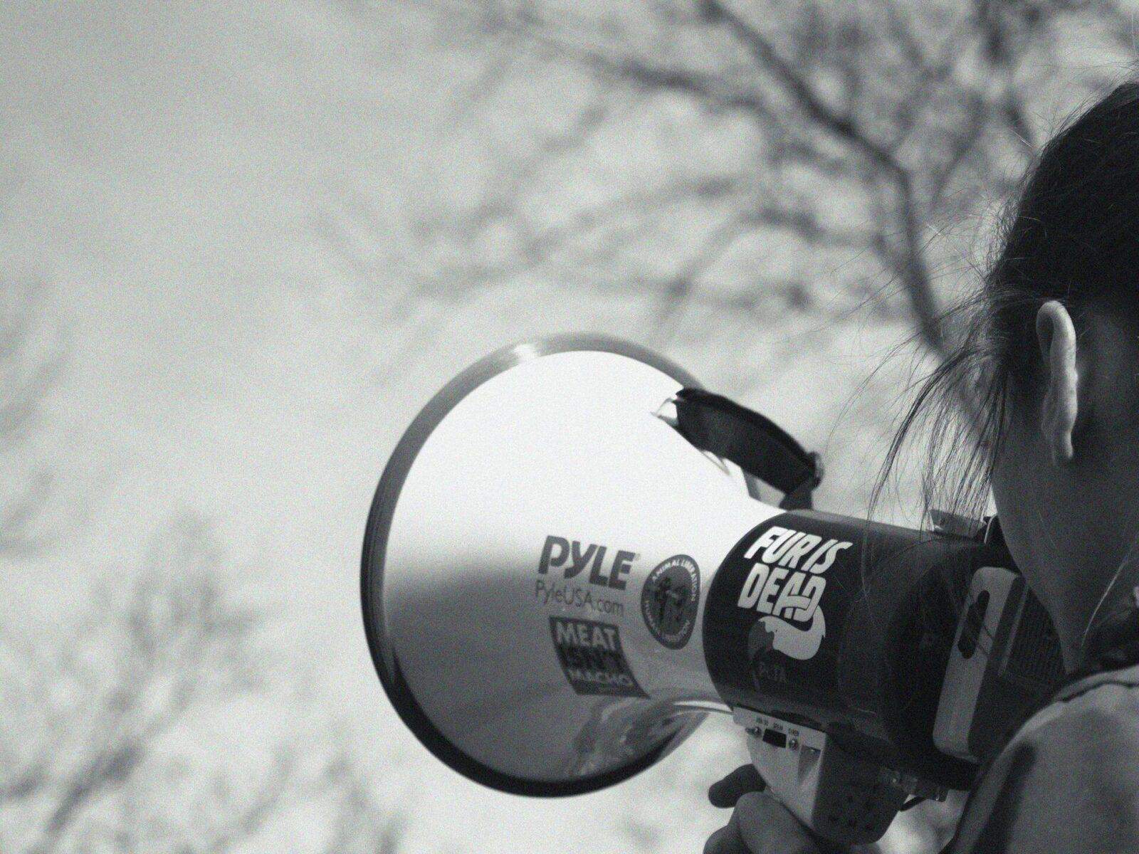 Black and white photo of a person holding a megaphone with the words "FUR IS DEAD" written on it.