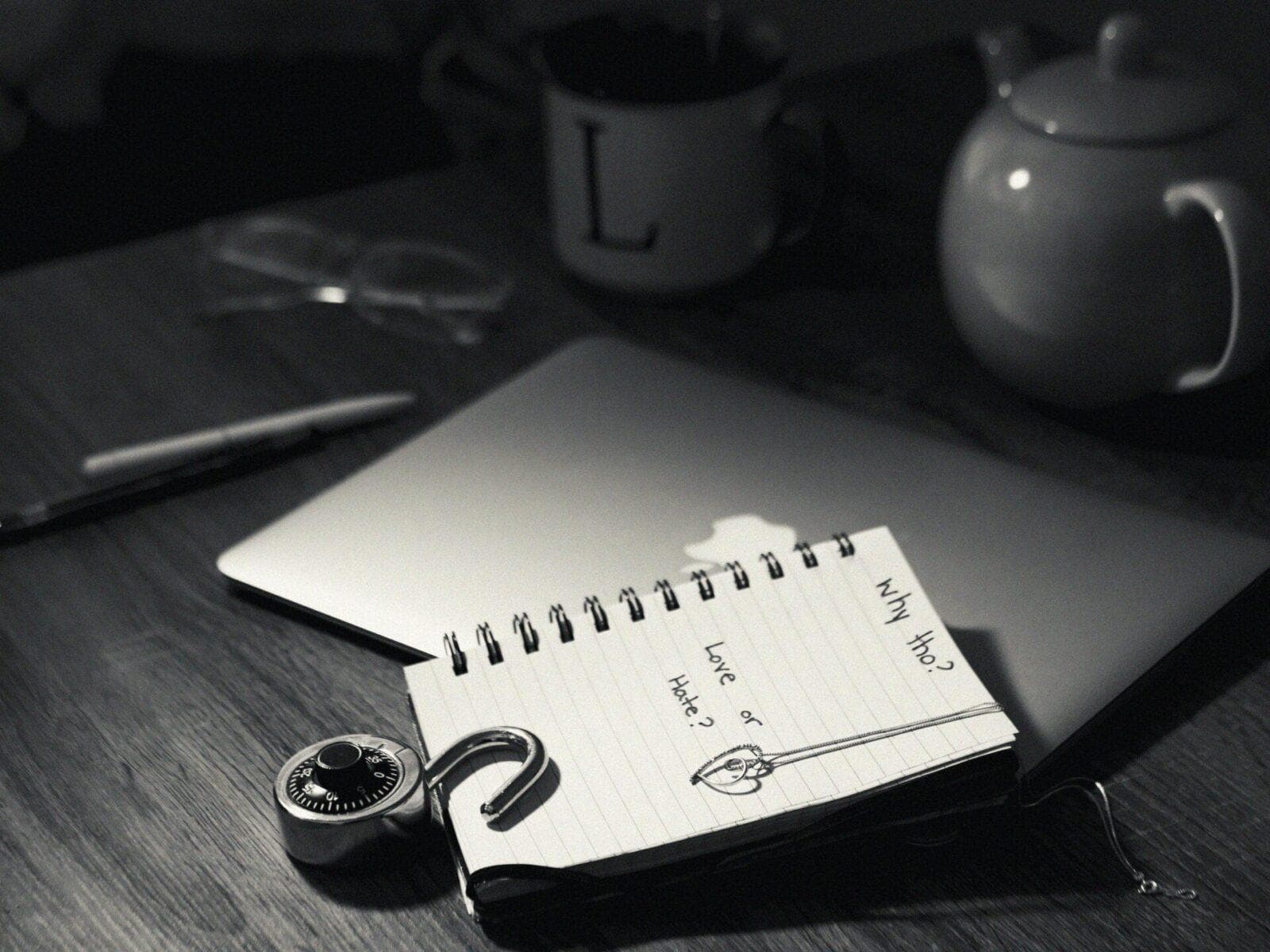 Black and white photo of a notebook with the words "Why Tho?" written on it, a pen, a padlock with a key, glasses, and a laptop on a wooden table.