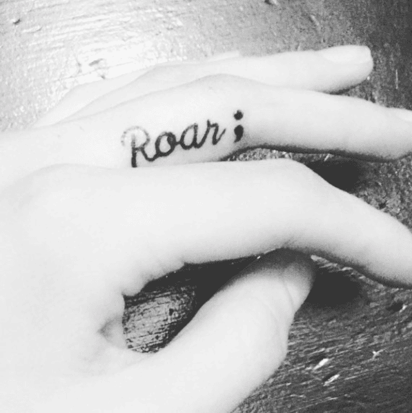 Black and white photograph of a person's finger with a tattoo of the word "Roar;" written in a cursive script.