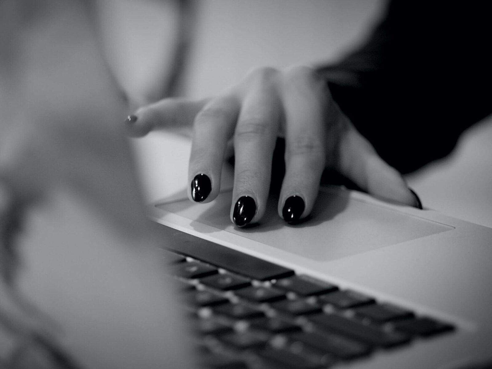 Black and white photo of a hand with dark nail polish using a laptop touchpad.