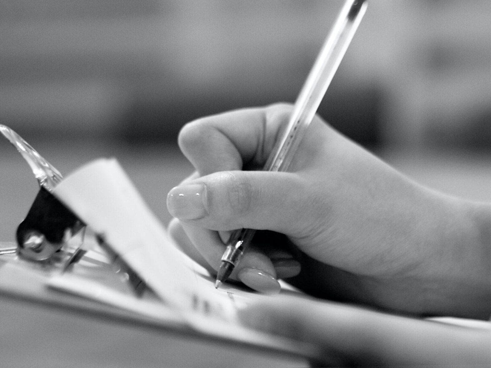 Black and white close-up photo of a hand writing on a clipboard with a pen.