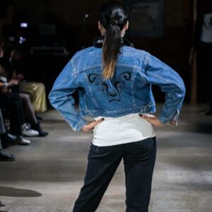 A model walks the runway at The Vegan Fashion Show wearing a stylish denim jacket featuring the Dandi Lion logo from The Honest Whisper, a vegan brand committed to ethical and sustainable fashion.