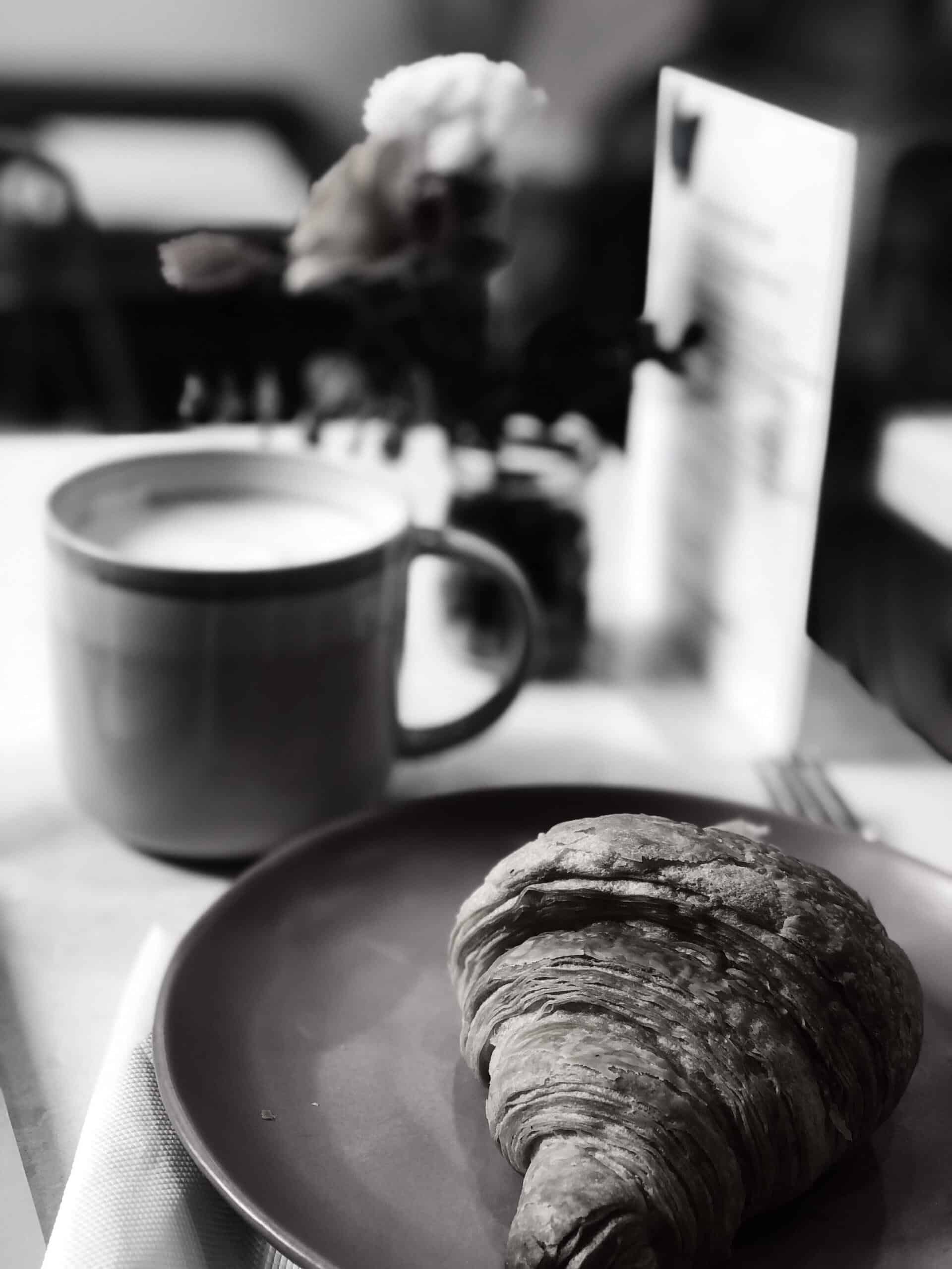 Black and white photo of a cup of coffee and a pastry on a table, evoking a cozy breakfast scene.