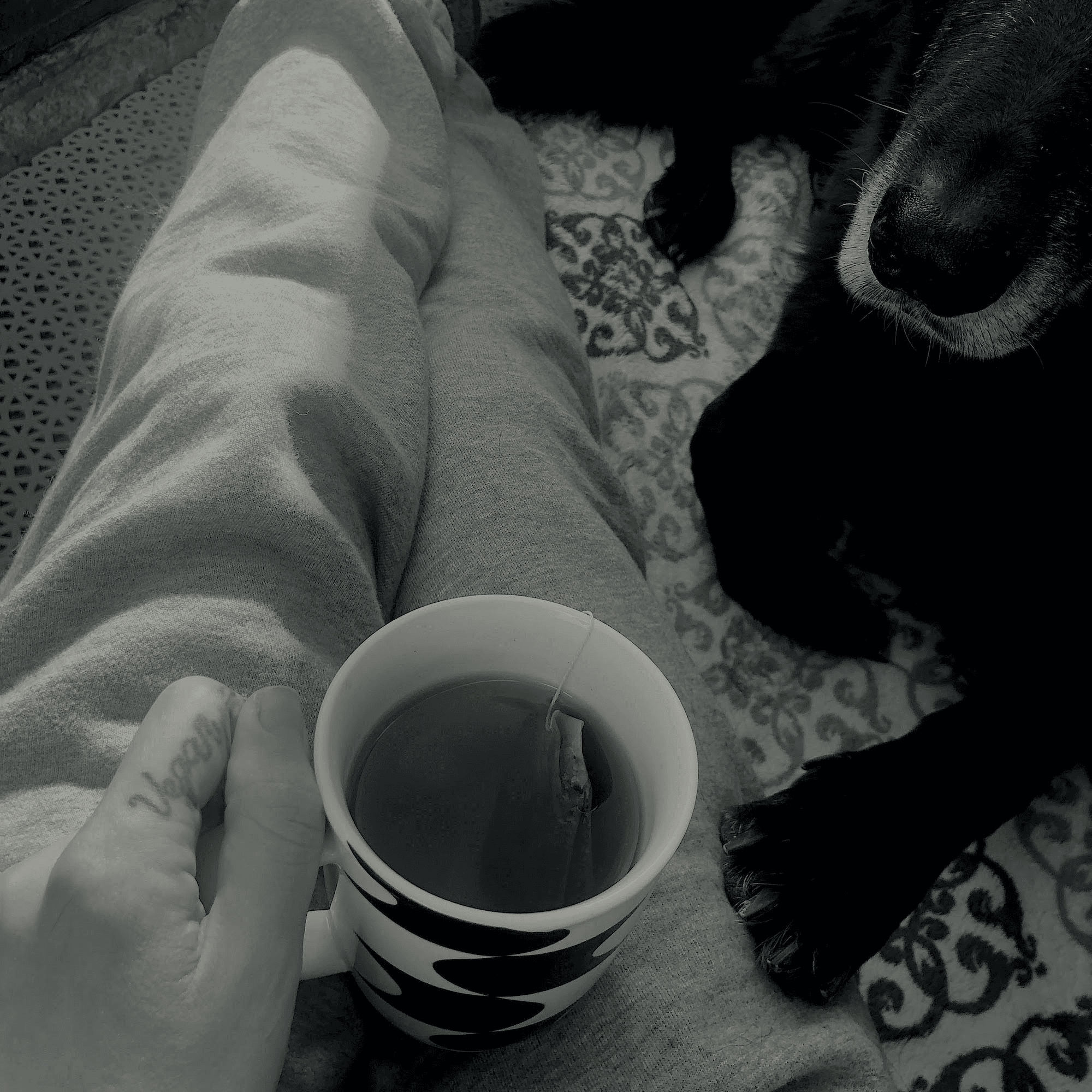 Black and white photo of a person holding a cup of tea, with a dog's paw visible in the foreground and a vegan tattoo on the person's finger.