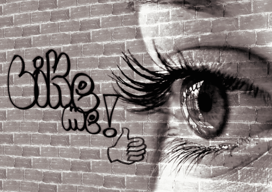A black and white image of an eye with the word "LIKE ME!" graffiti-style on a brick wall, symbolizing the pressure of social media.