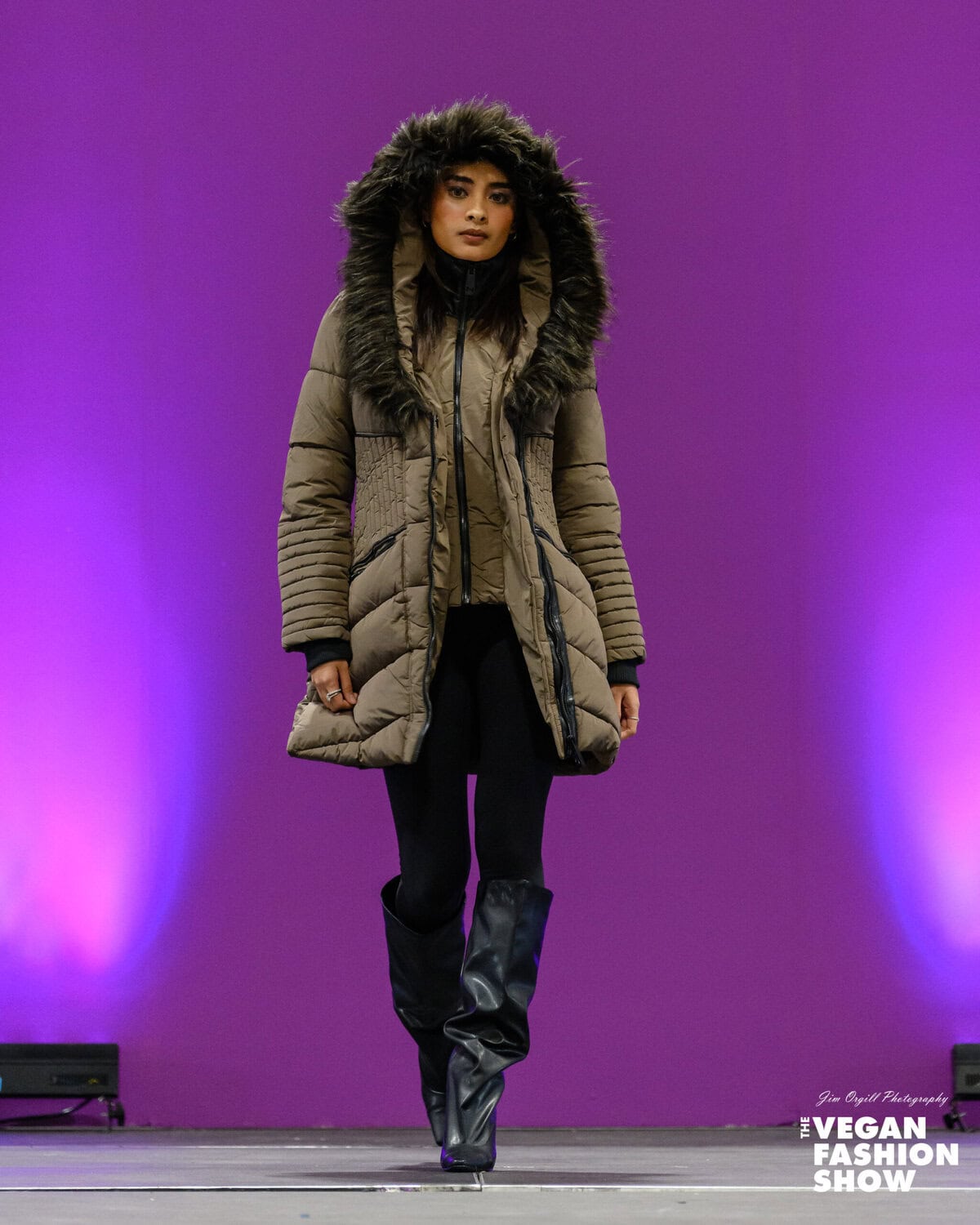 Warm winter coat with fur hood on runway Model Naomi (naomicabrera.__) walks the runway in a full-length, brown, quilted Gigi Mid Length Parka with a faux-fur hood by NOIZE, paired with black pants and knee-high boots, at The Vegan Fashion Show.